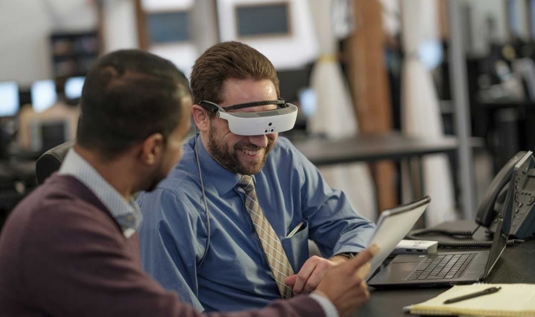 Two men look at a document while one of them tests out the new eSight wearable technology glasses