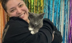 Maricella smiling as she holds gray kitten Shadow in front of a colorful backdrop.
